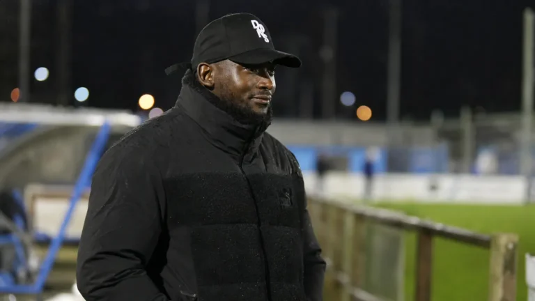 George Desmond Kamurasi aan de zijlijn van een Engels amateurvoetbalveld in donkere bovenkleding.