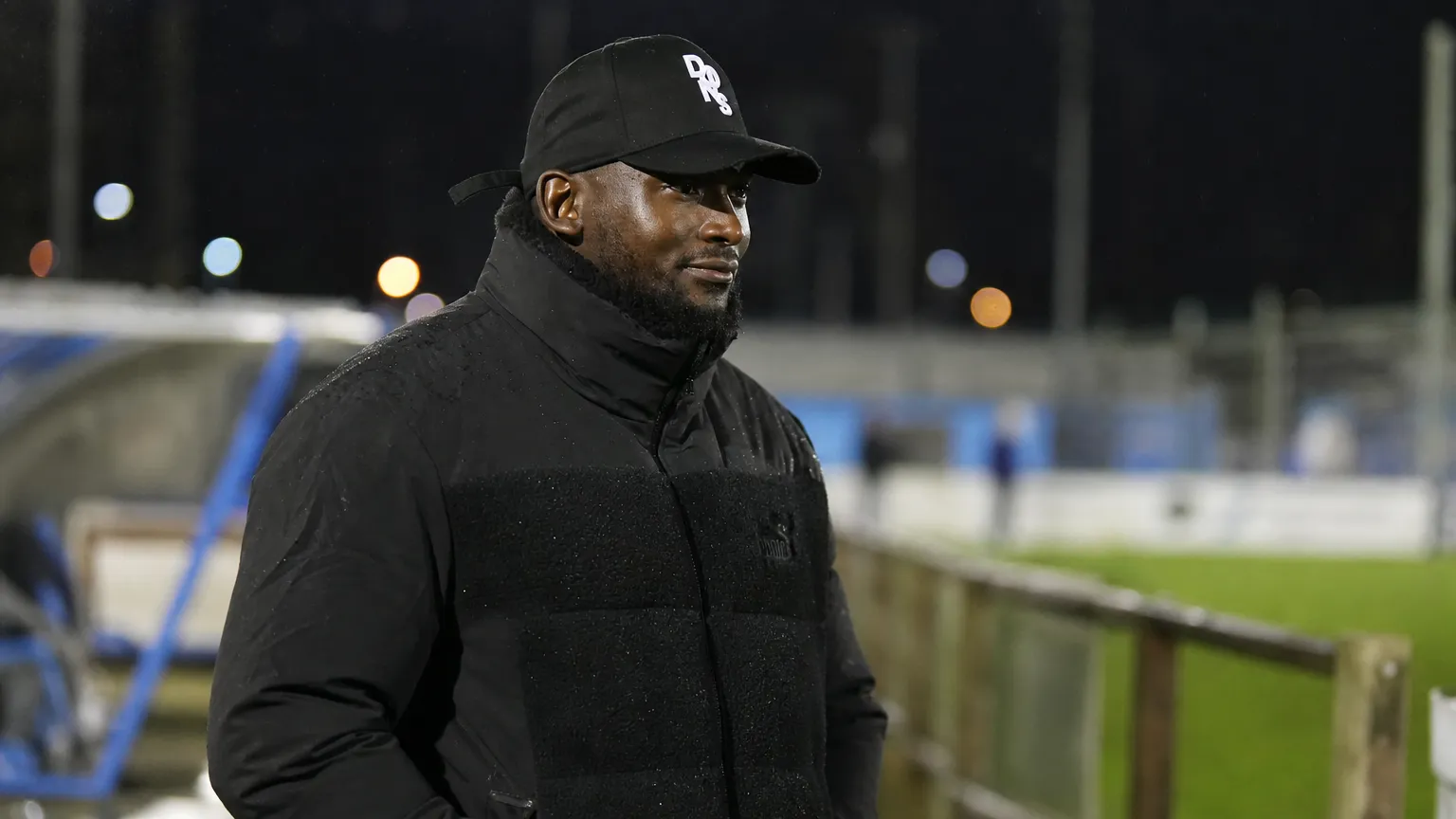 George Desmond Kamurasi aan de zijlijn van een Engels amateurvoetbalveld in donkere bovenkleding.