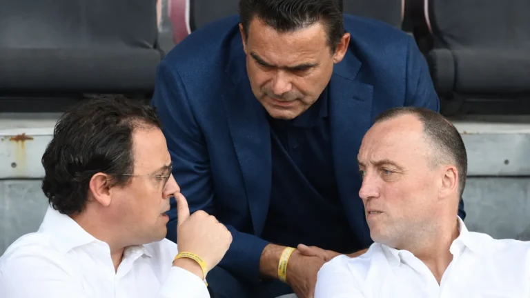 Drie niet-spelende clubvertegenwoordigers in gesprek op de tribune van een stadion.