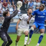 Voetballers in blauwe en witte tenues strijden voor doel terwijl een doelman uitkomt.