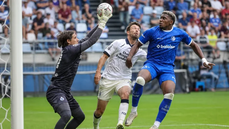 Voetballers in blauwe en witte tenues strijden voor doel terwijl een doelman uitkomt.