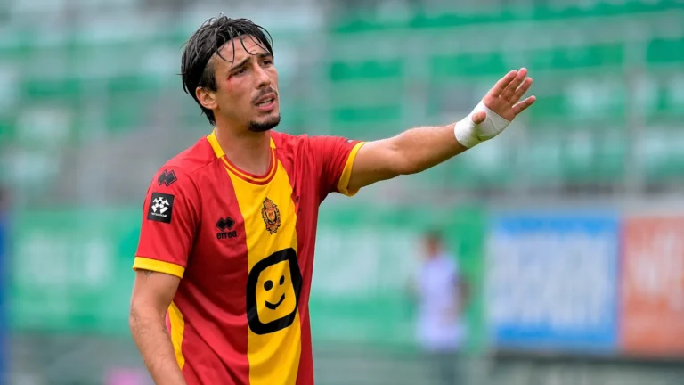 Een voetballer in een rood-geel shirt gebaart langs de rand van het veld.