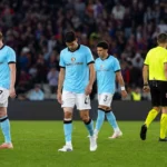 Spelers en een scheidsrechter op het veld na een wedstrijd, met supporters op de tribune.