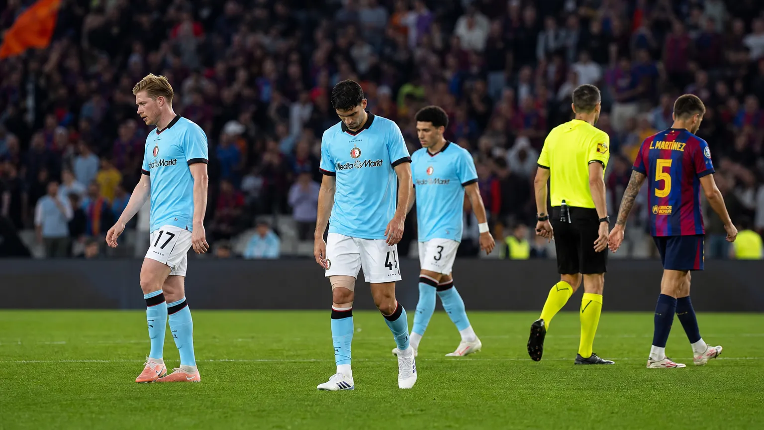 Spelers en een scheidsrechter op het veld na een wedstrijd, met supporters op de tribune.