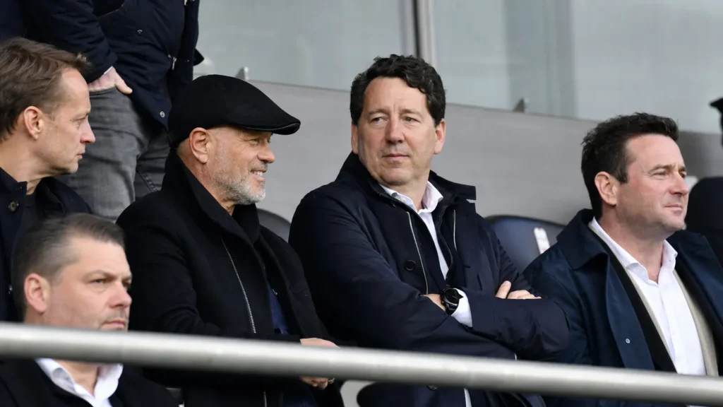 Niet-spelende clubofficials in donkere jassen in een stadiontribune.