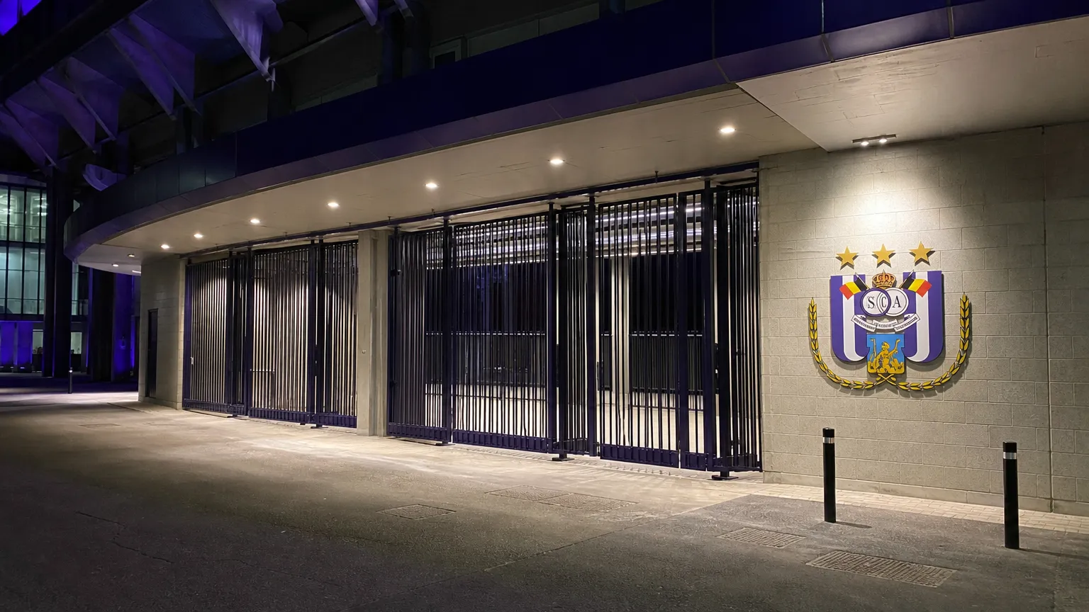 Verlichte ingang van Lotto Park met één Anderlecht-embleem aan de gevel.