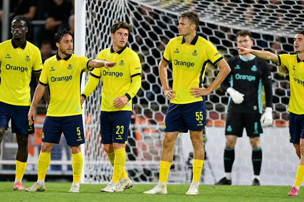 Een groep voetballers in gele shirts vormt een verdedigende muur voor het doel.