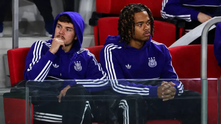 Een groep Anderlecht-spelers in paarse trainingskleding kijkt vanuit de stadionzitjes toe.