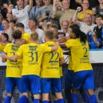 Een groep voetballers in gele shirts viert samen voor een volle thuistribune.