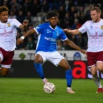 Een voetballer in blauw schermt de bal af terwijl twee tegenstanders hem achtervolgen.