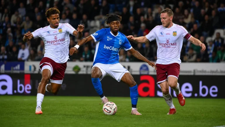Een voetballer in blauw schermt de bal af terwijl twee tegenstanders hem achtervolgen.