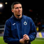 Hans Vanaken in een donkerblauwe sportjas naast een voetbalveld.