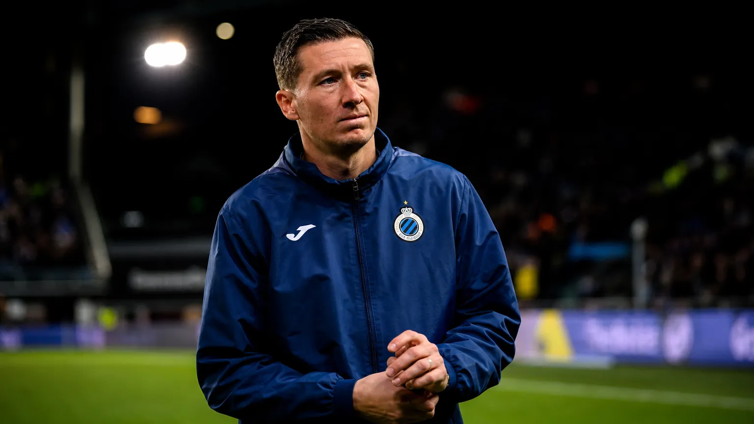 Hans Vanaken in een donkerblauwe sportjas naast een voetbalveld.