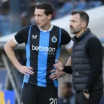 Een Club Brugge-speler in blauw-zwart overlegt langs het veld met een staflid.