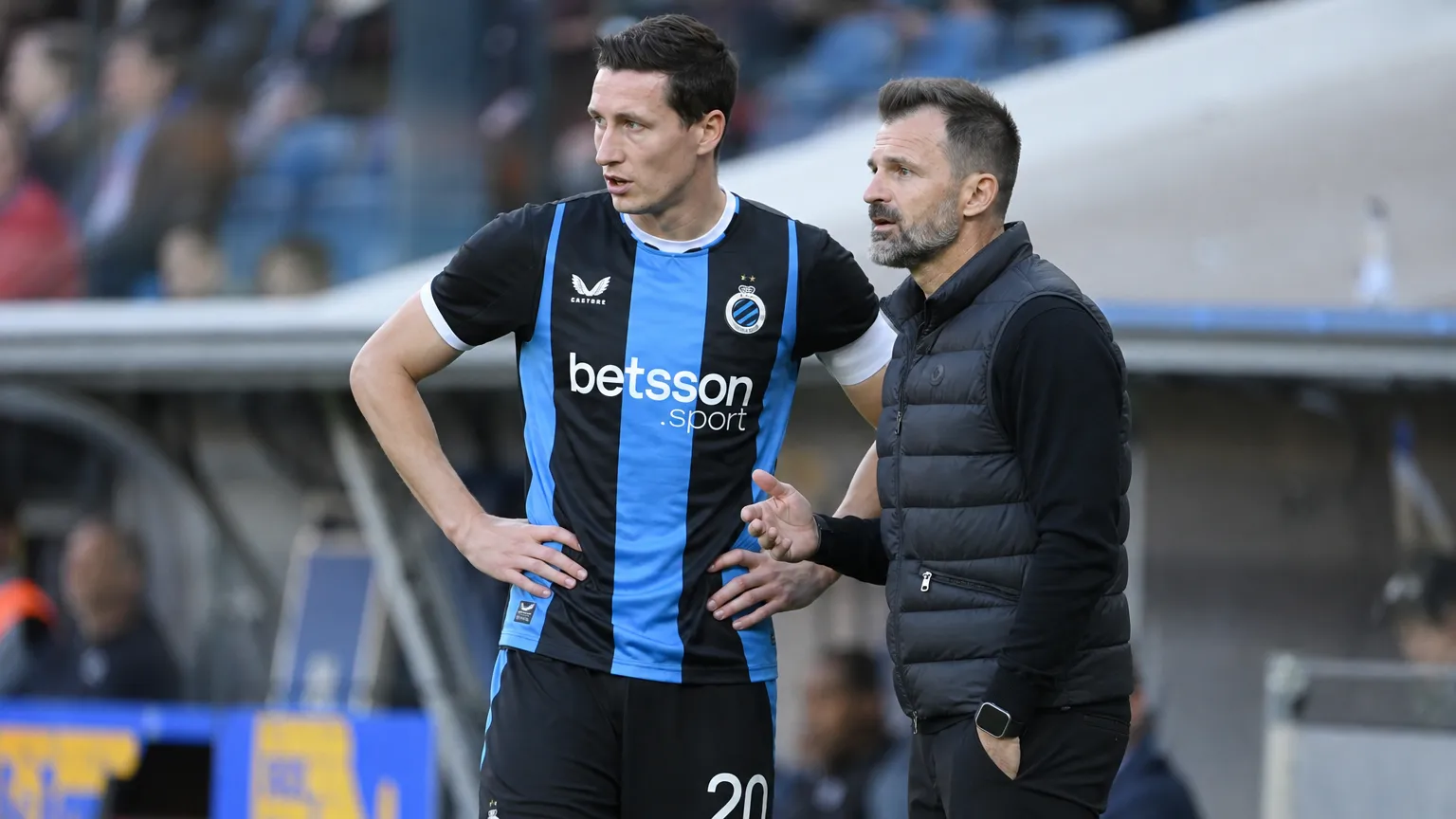 Een Club Brugge-speler in blauw-zwart overlegt langs het veld met een staflid.