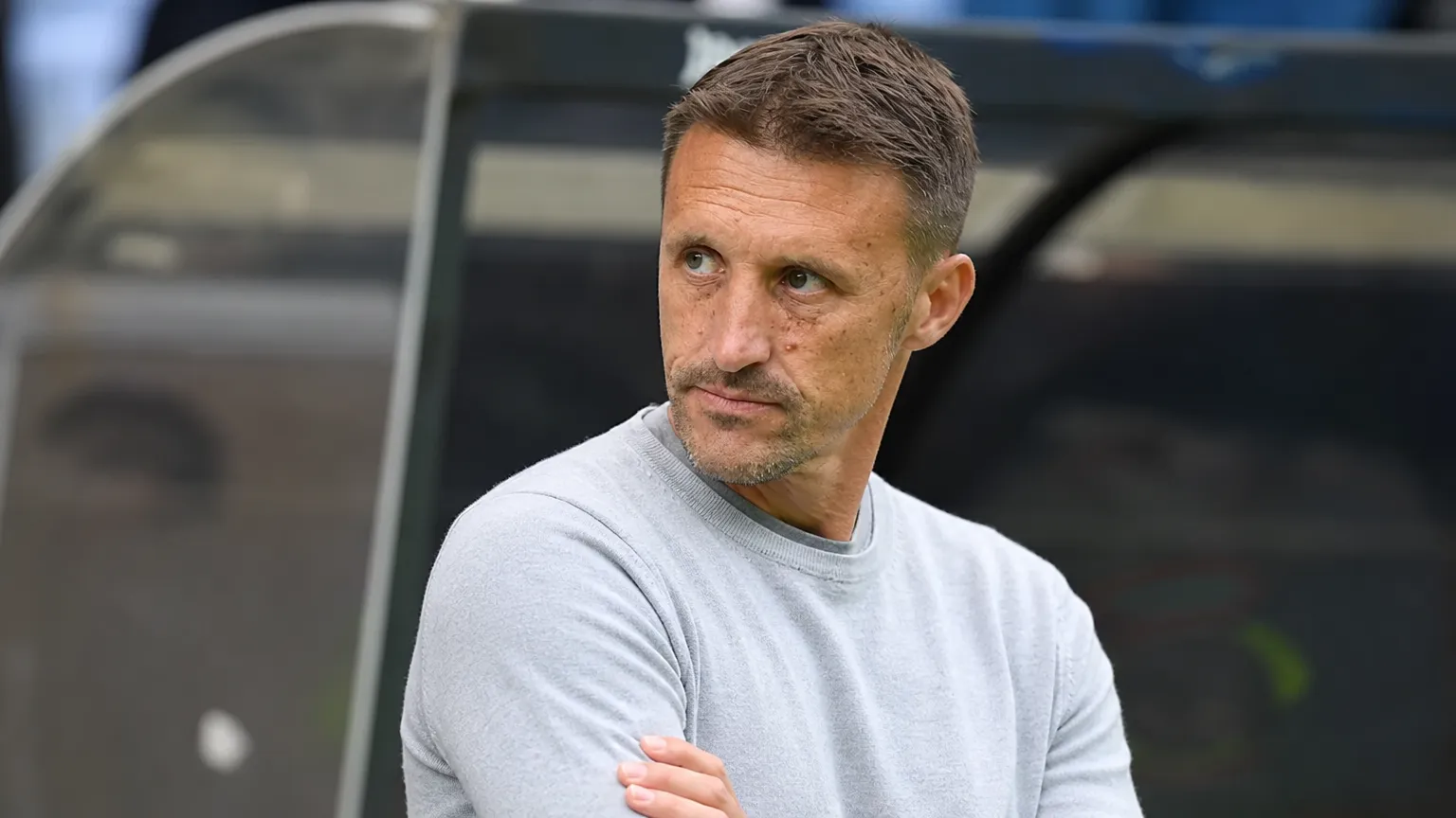 Een staflid in een lichtgrijze trui kijkt geconcentreerd vanaf de technische zone naar het veld.