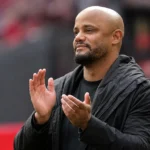 Een coach in donkere jas applaudisseert langs de zijlijn van een voetbalstadion.