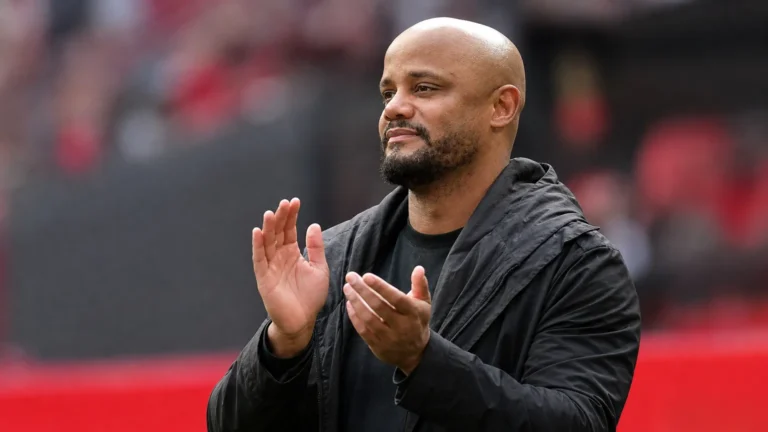 Een coach in donkere jas applaudisseert langs de zijlijn van een voetbalstadion.