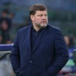 Een man in een donkerblauwe jas observeert langs de zijlijn een voetbalwedstrijd.