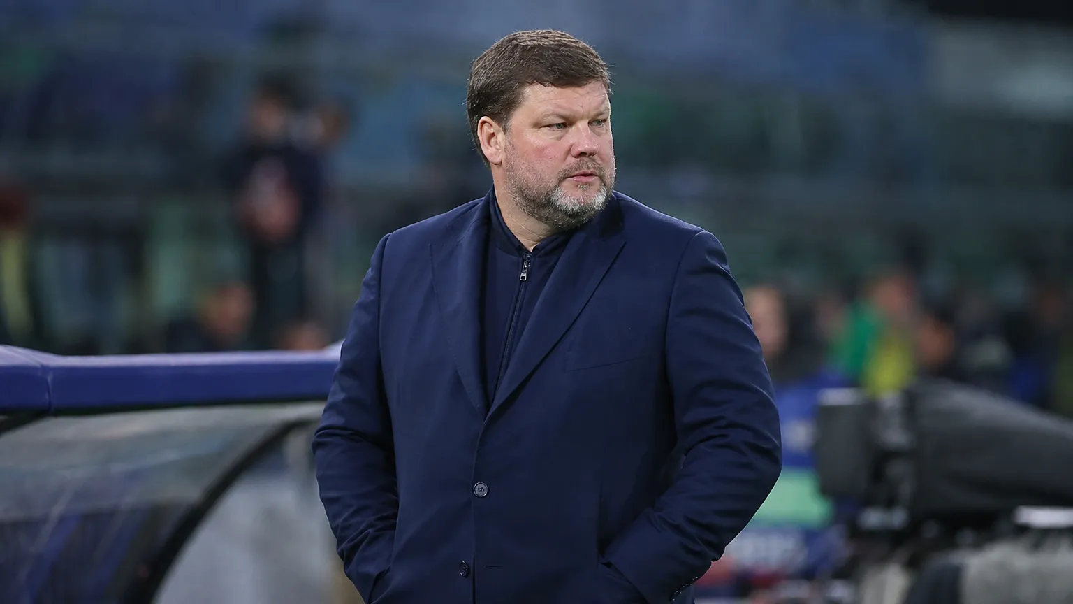 Een man in een donkerblauwe jas observeert langs de zijlijn een voetbalwedstrijd.