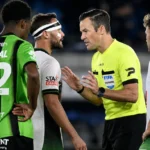 Een scheidsrechter bespreekt een fase met spelers in groene en witte wedstrijdtenues.