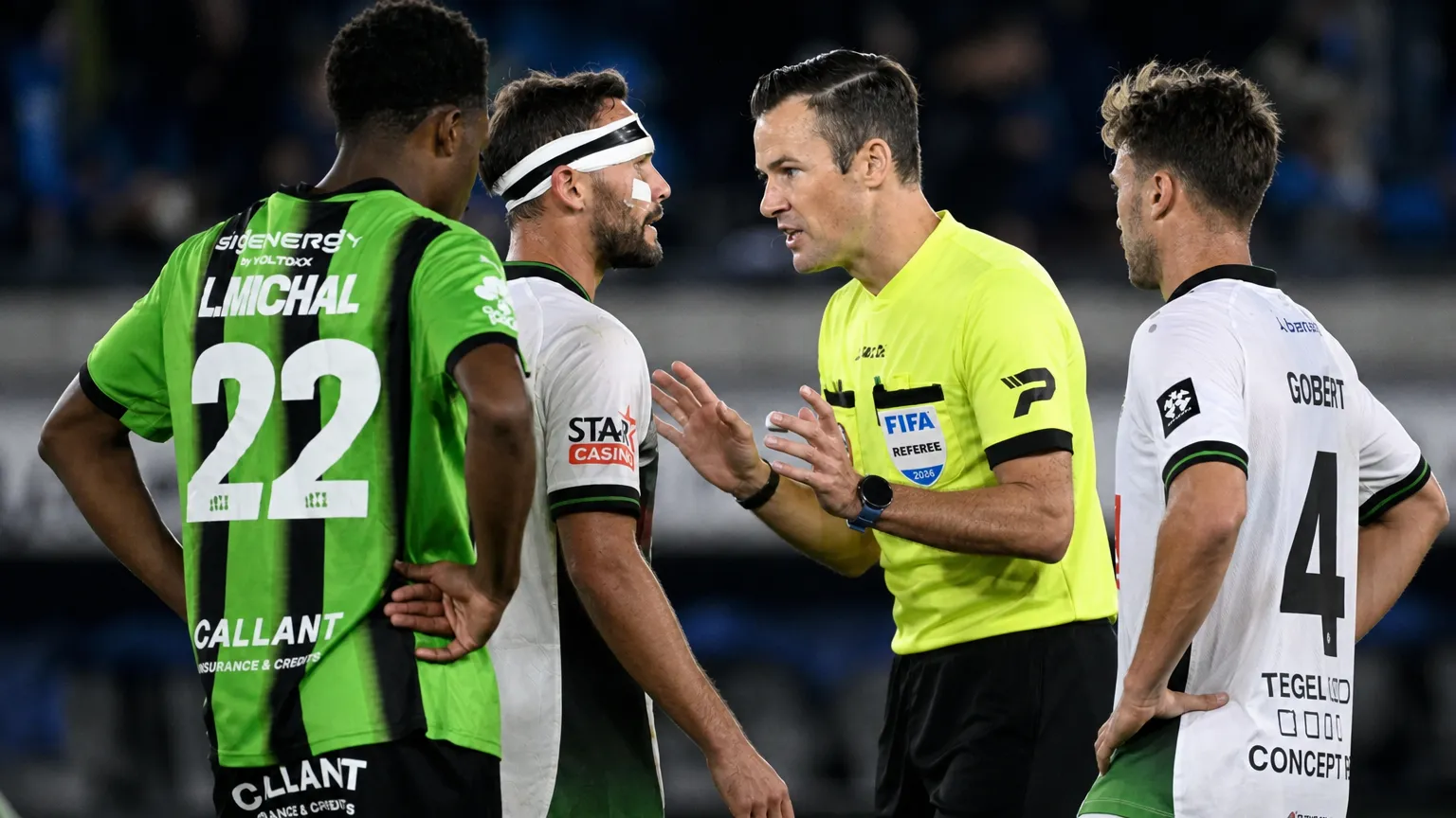 Een scheidsrechter bespreekt een fase met spelers in groene en witte wedstrijdtenues.