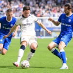 Voetballers in witte en blauwe tenues vechten om de bal op een zonnig veld.