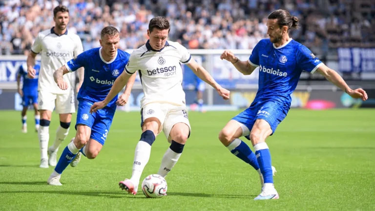 Voetballers in witte en blauwe tenues vechten om de bal op een zonnig veld.