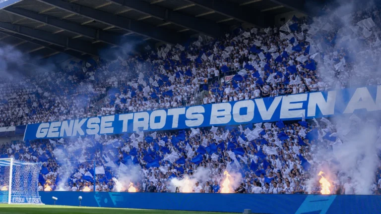 Collectieve KRC Genk-supporters met blauw-witte vlaggen op de thuistribune voor de aftrap.