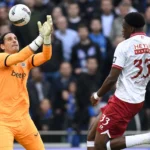 Een doelman en een veldspeler in actie in het strafschopgebied tijdens een Belgische clubwedstrijd.