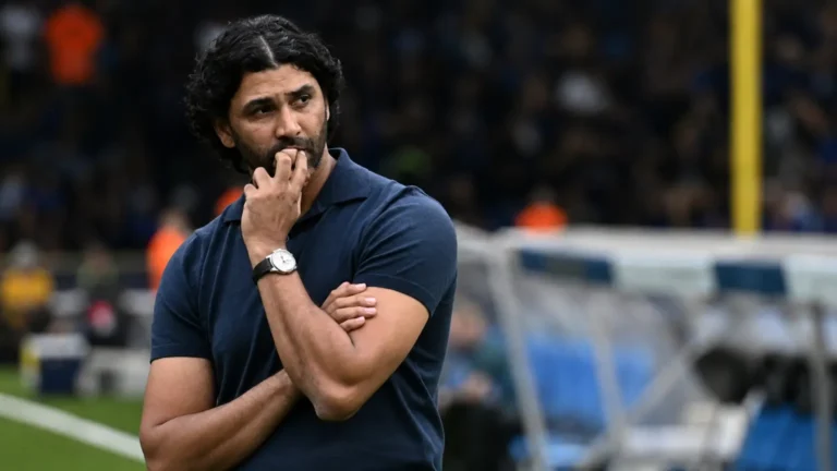 Een coach in een donkerblauwe polo staat bedachtzaam aan de zijlijn van een stadion.