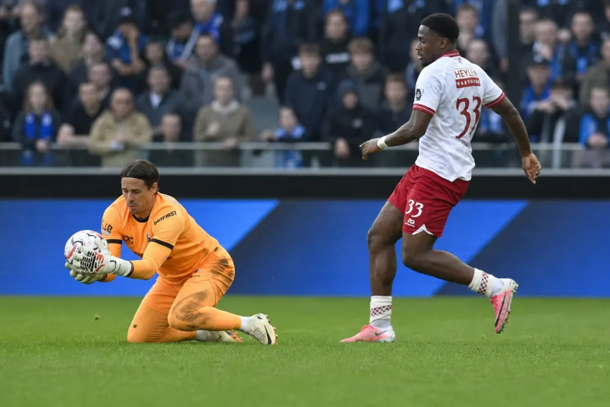 Een doelman in oranje vangt een lage bal terwijl een tegenstander afremt op het veld.