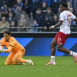 Een doelman in oranje vangt een lage bal terwijl een tegenstander afremt op het veld.