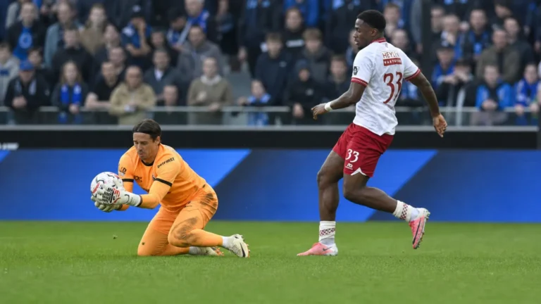 Een doelman in oranje vangt een lage bal terwijl een tegenstander afremt op het veld.