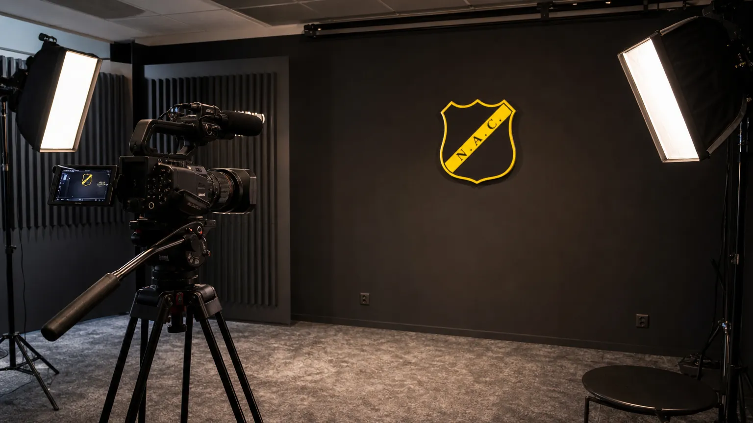 Voorbereide clubmediastudio van NAC Breda met camera-apparatuur en een NAC-embleem aan de wand.