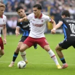 Voetballers in witte en donkere tenues tijdens een duel op het veld.