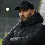 Een coach van Patro Eisden kijkt aandachtig langs de zijlijn in de regen.