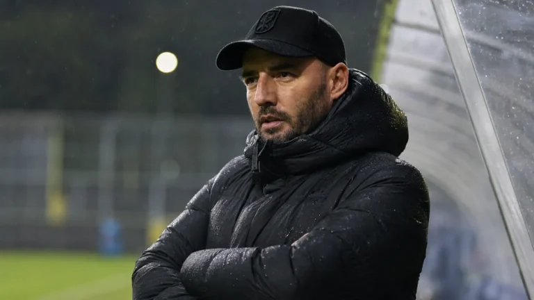 Een coach van Patro Eisden kijkt aandachtig langs de zijlijn in de regen.