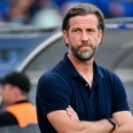 Een coach in donkerblauwe polo kijkt geconcentreerd vanuit de technische zone van een voetbalstadion.