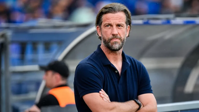 Een coach in donkerblauwe polo kijkt geconcentreerd vanuit de technische zone van een voetbalstadion.