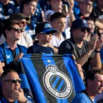 Een groep Club Brugge-supporters in blauw-zwarte kledij op een zonnige tribune.