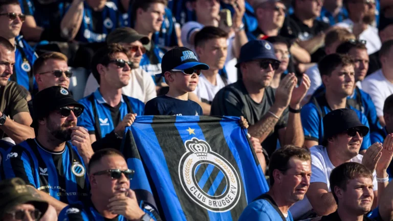 Een groep Club Brugge-supporters in blauw-zwarte kledij op een zonnige tribune.
