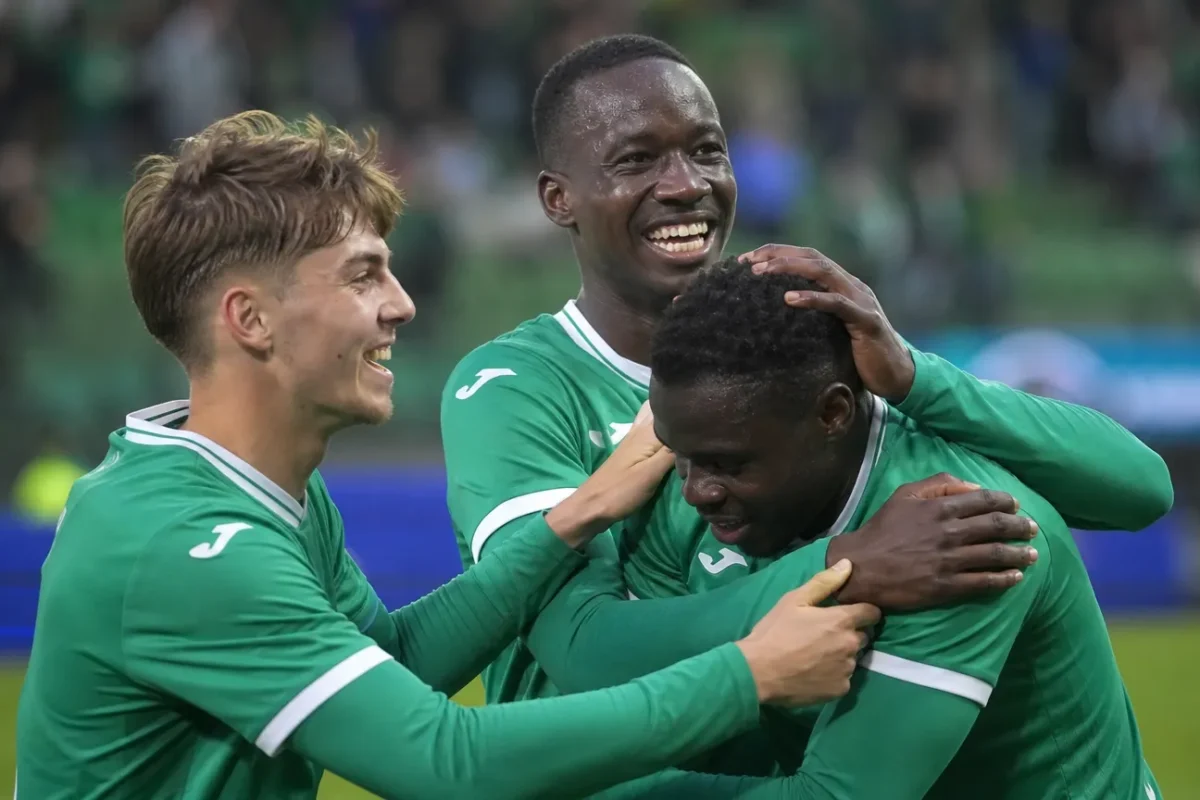 Drie voetballers in groene shirts vieren samen een doelpunt op het veld.
