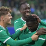 Drie voetballers in groene shirts vieren samen een doelpunt op het veld.