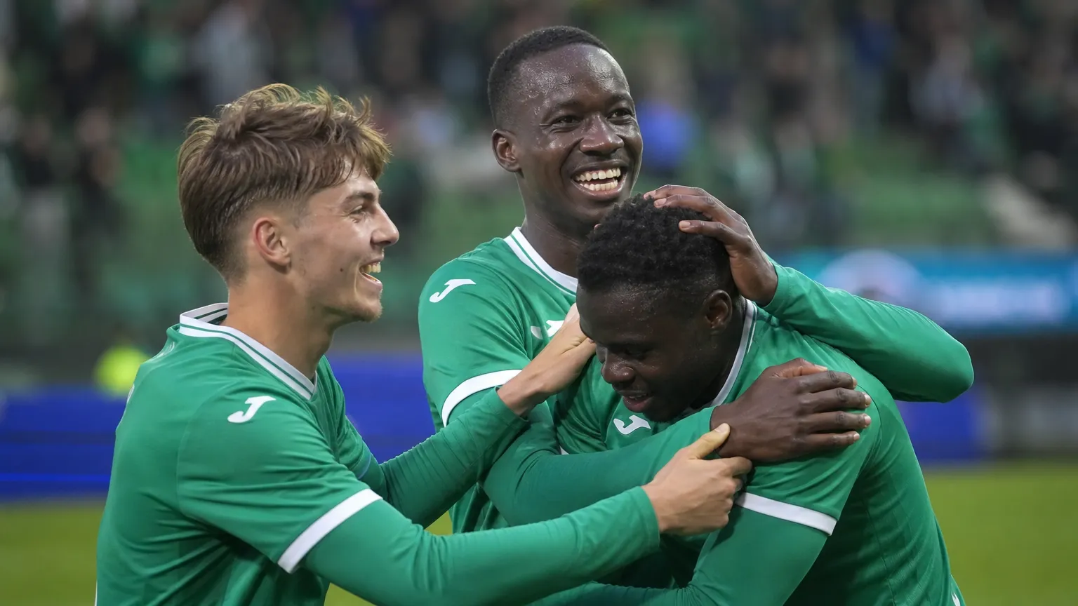 Drie voetballers in groene shirts vieren samen een doelpunt op het veld.