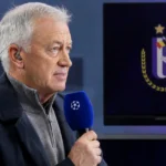 Een voetbalanalist met microfoon in een televisiestudio met een subtiel RSC Anderlecht-embleem op een vaste monitor.