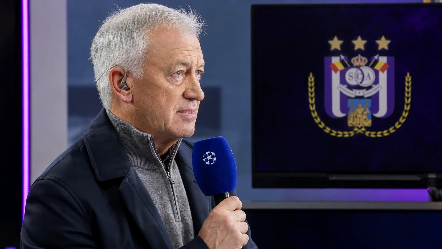 Een voetbalanalist met microfoon in een televisiestudio met een subtiel RSC Anderlecht-embleem op een vaste monitor.