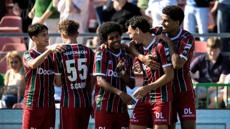 Een groep voetballers in rood-groen-wit gestreepte shirts viert samen naast de tribune.
