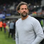 Een niet-spelende clubmedewerker in een grijze trui aan de zijlijn van een voetbalveld.