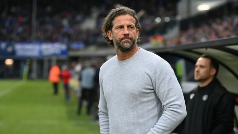 Een niet-spelende clubmedewerker in een grijze trui aan de zijlijn van een voetbalveld.